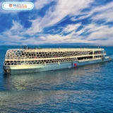 A large, modern, white mega yacht with a distinctive hexagonal pattern on its upper deck, sailing on blue water under a cloudy sky. The Mayra Tours logo is visible at the top.